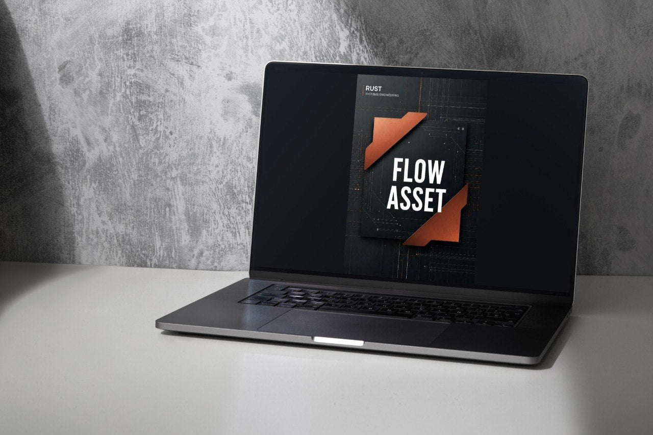 A dark gray laptop sits open on a white surface against a textured gray wall, with the screen displaying a dark graphic that reads 'FLOW ASSET'.